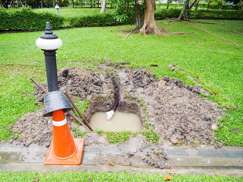 Sewer line excavation site with orange traffic cone and water pooling in a muddy trench, indicating potential plumbing issues and the need for professional repair services.
