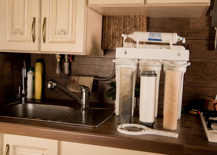 Whole-house water filtration system installed under kitchen sink, featuring multiple filter cartridges and connected plumbing, emphasizing clean water solutions for home improvement.